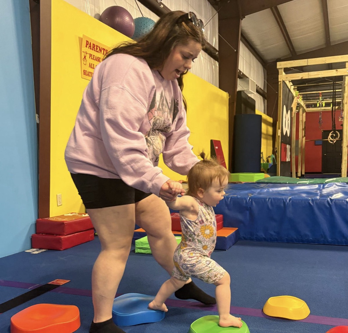 A parent holds a toddler's hand as she steps across colored stepping stones in a Playful Parenting class