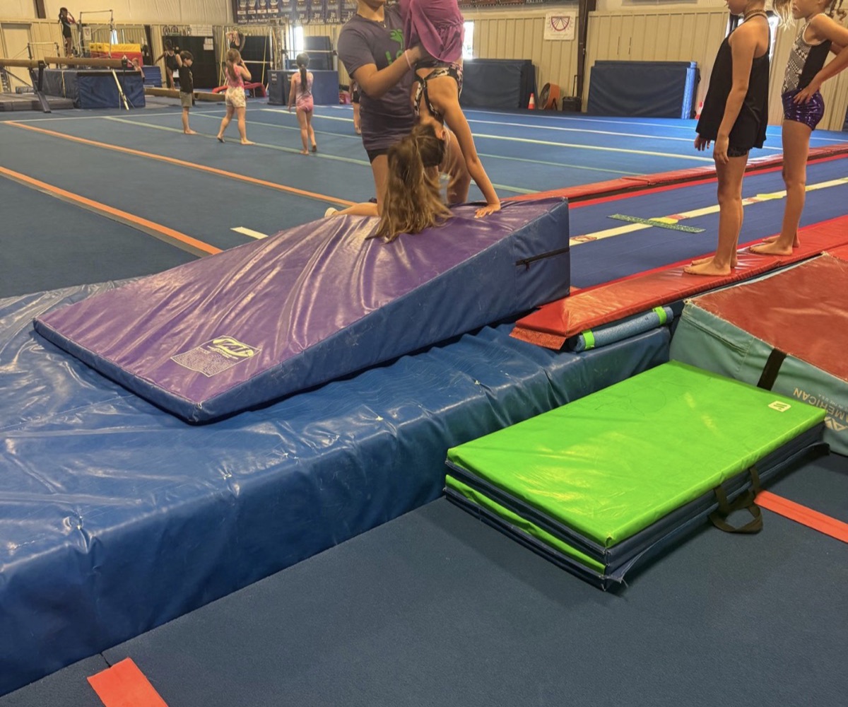 A coach spots a gymnast in a handstand on an incline mat