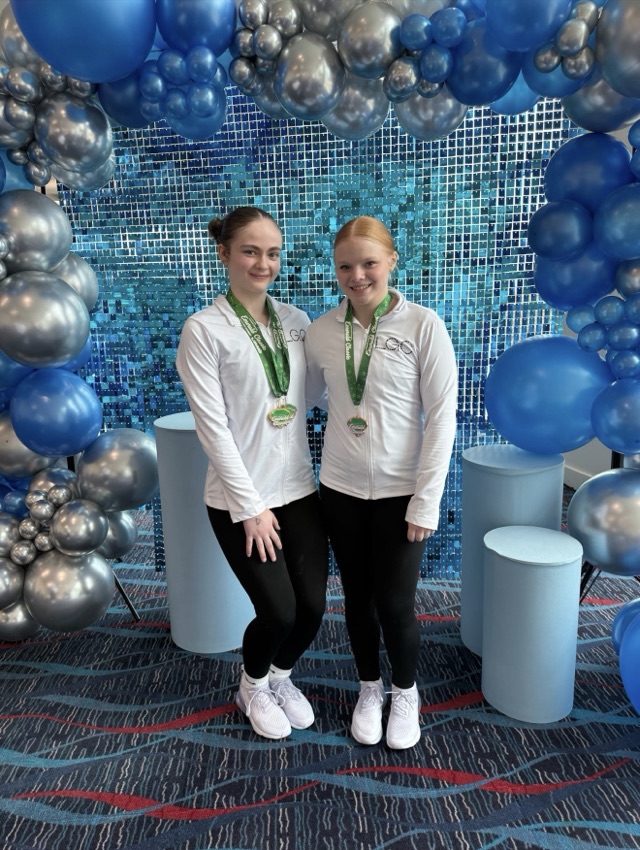 Two team gymnasts in white LGC warm-up jackets with medals in front of a balloon arch