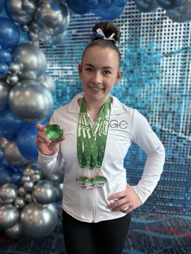 A team gymnast holding an Emerald Classic gem trophy and wearing three medals
