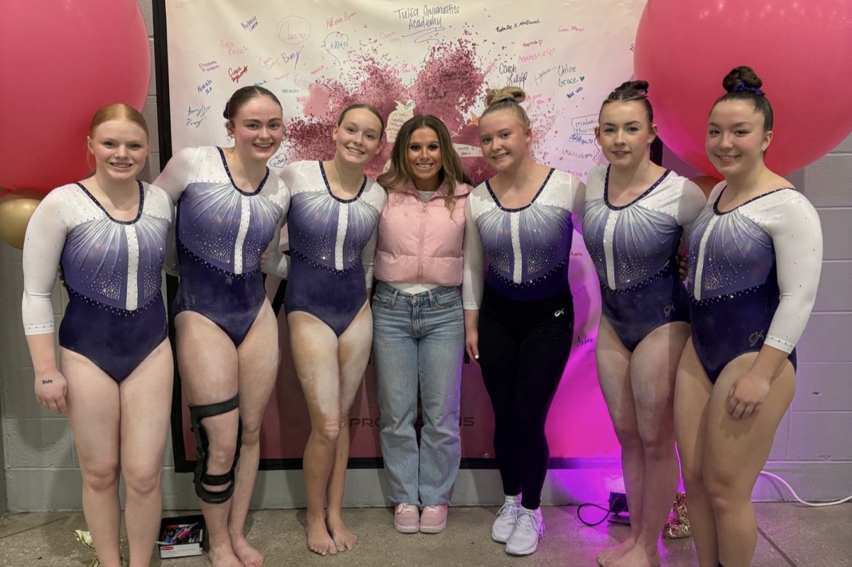 Six team gymnasts and a coach pose in front of a pink signature banner
