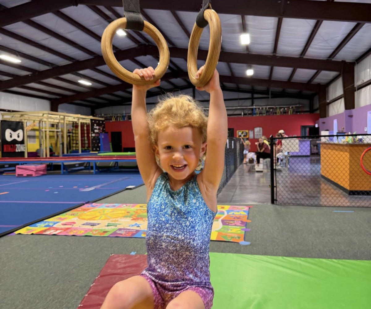 A Tiny Tots gymnast smiling while hanging from the rings at Leah's Gymnastics & Cheerleading